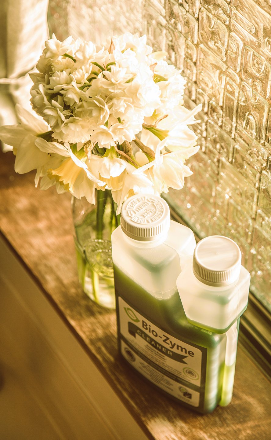 In front of a class window, Bio-Zyme Cleaner next to a jar with white flowers.